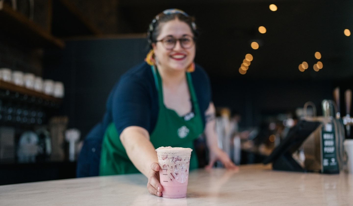 A Starbucks partner in green apron placing down a pink beverage on a countertop in a Starbucks store