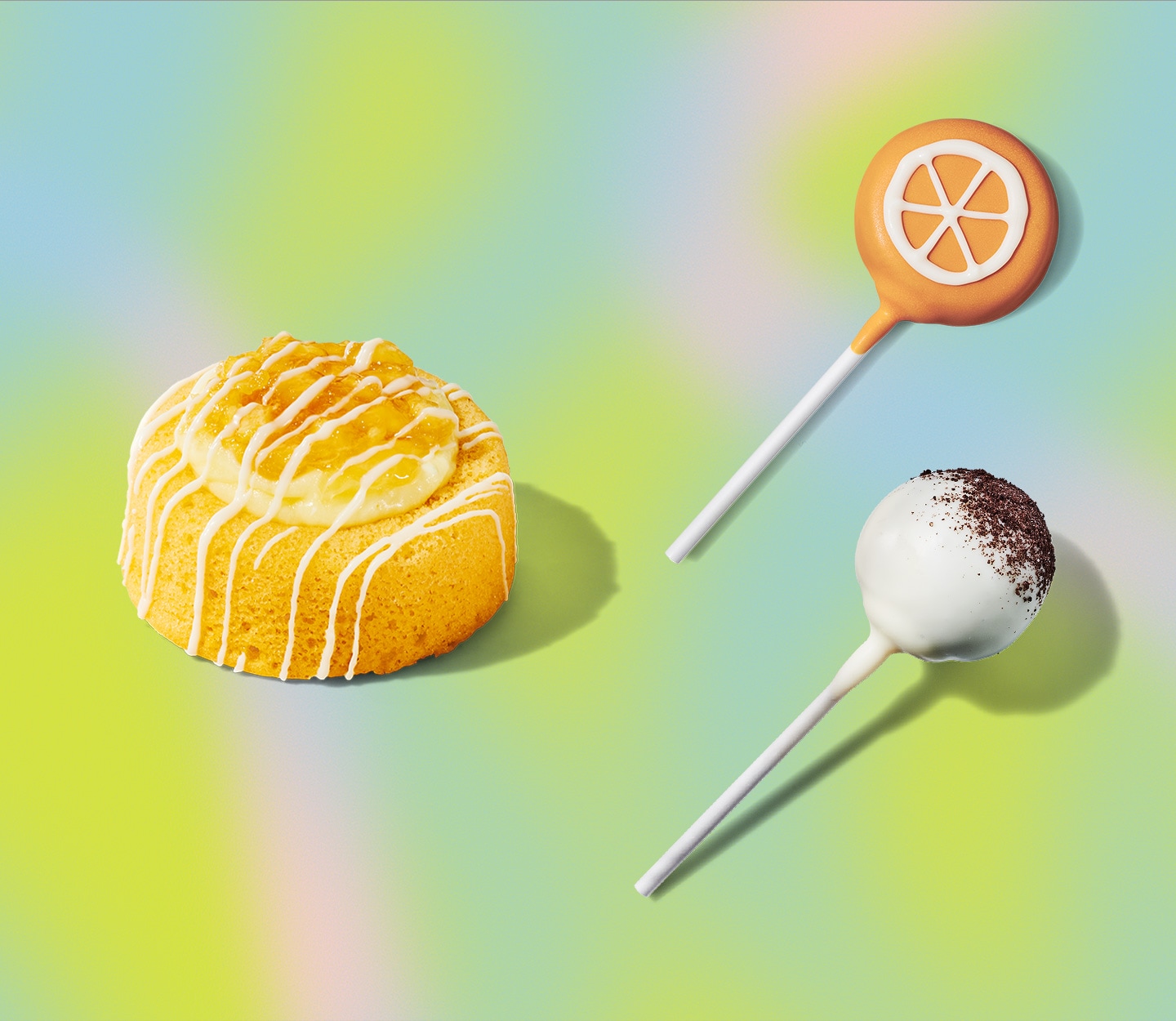 A round yellow cake with icing, an orange cake pop with orange slices drawn in icing, a white cake pop with chocolate cookie crumbs.