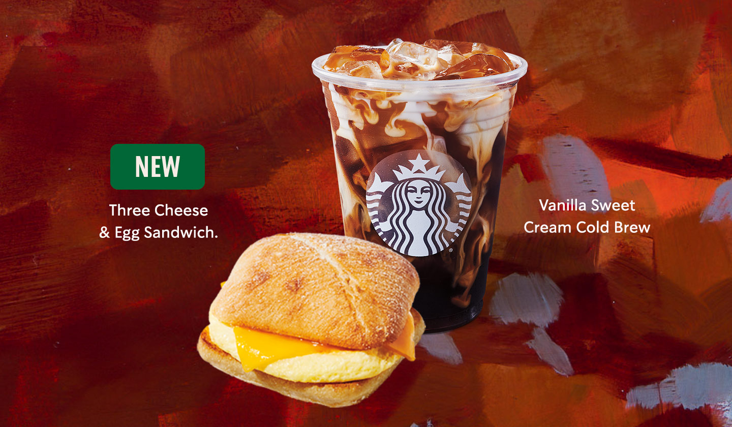 A fluffy, toasty, warm Three Cheese & Egg Sandwich, beside a Vanilla Sweet Cream Cold Brew marbling with its deep brown cold brew and fresh white cream