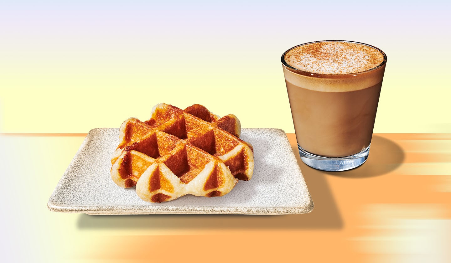 A waffle on a rectangular plate with a coffee based drink in a glass to its right, on a yellow and orange background.