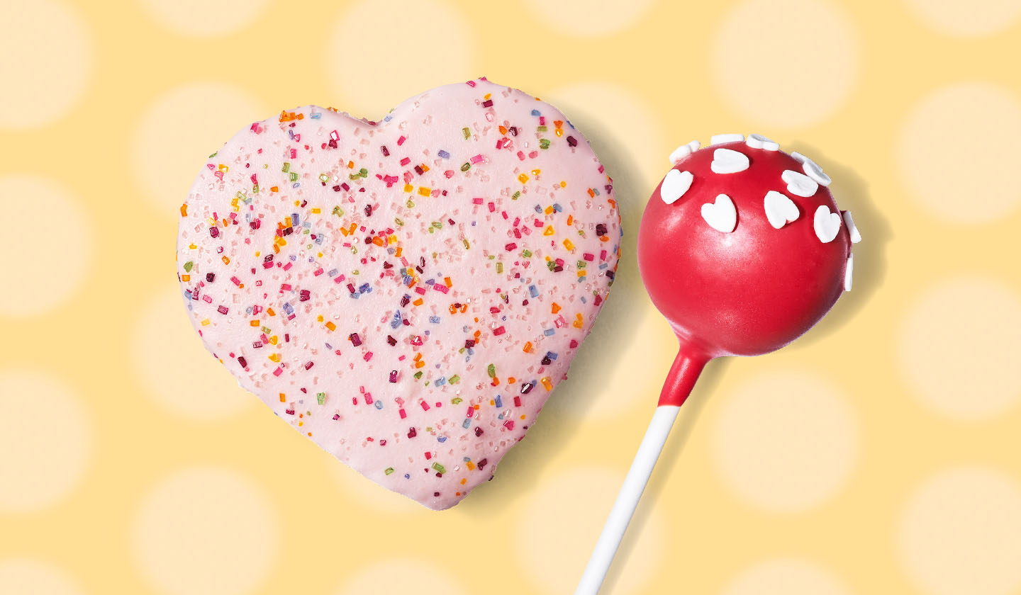 A heart shaped cookie with pink frosting and sprinkles and a red cake pop with white heart sprinkles on a yellow background