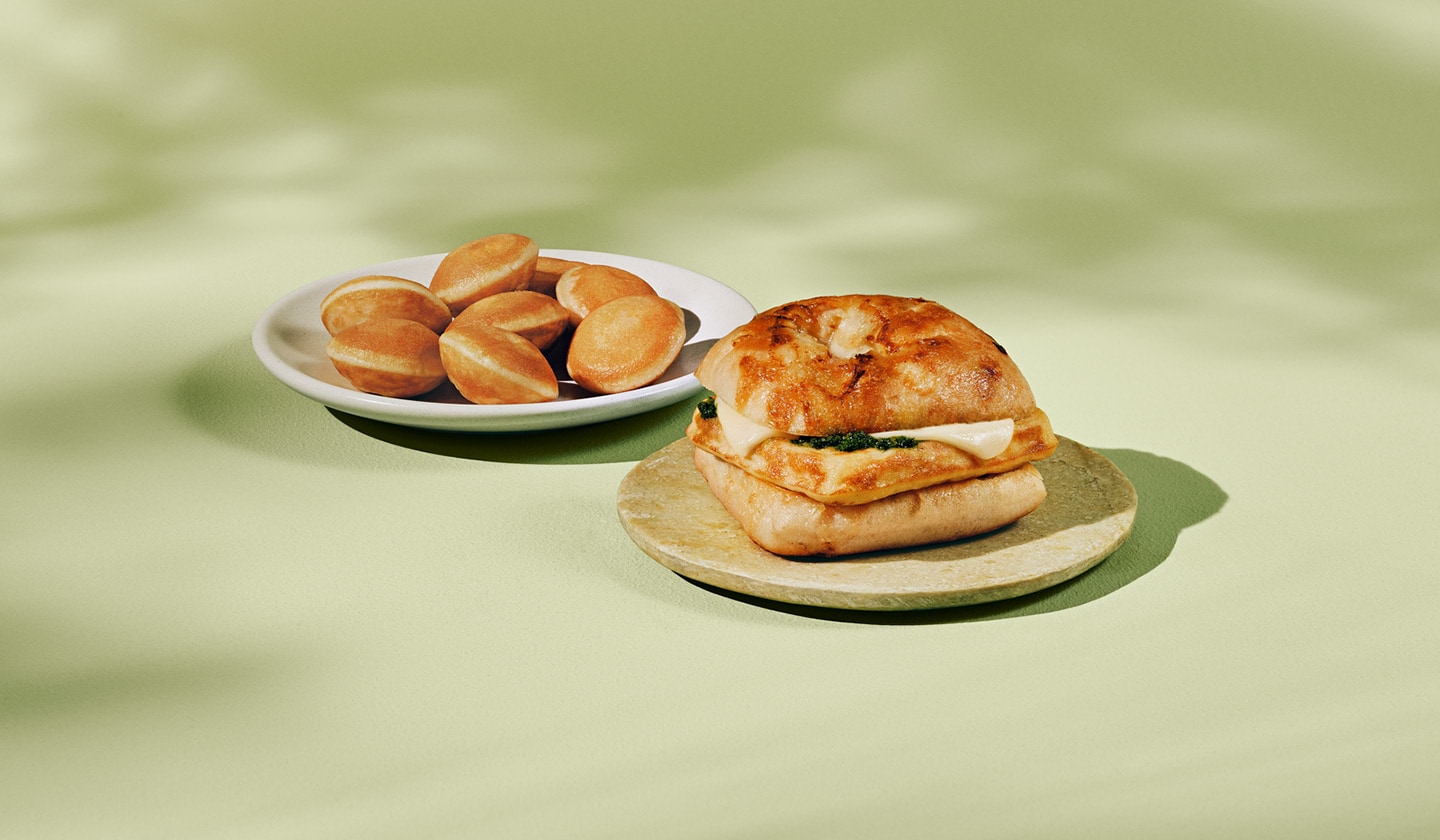 A light, fluffy, egg sandwich with melted cheese and pesto on a small plate, next to a plate of golden-brown mini pancakes, set on a light green background.