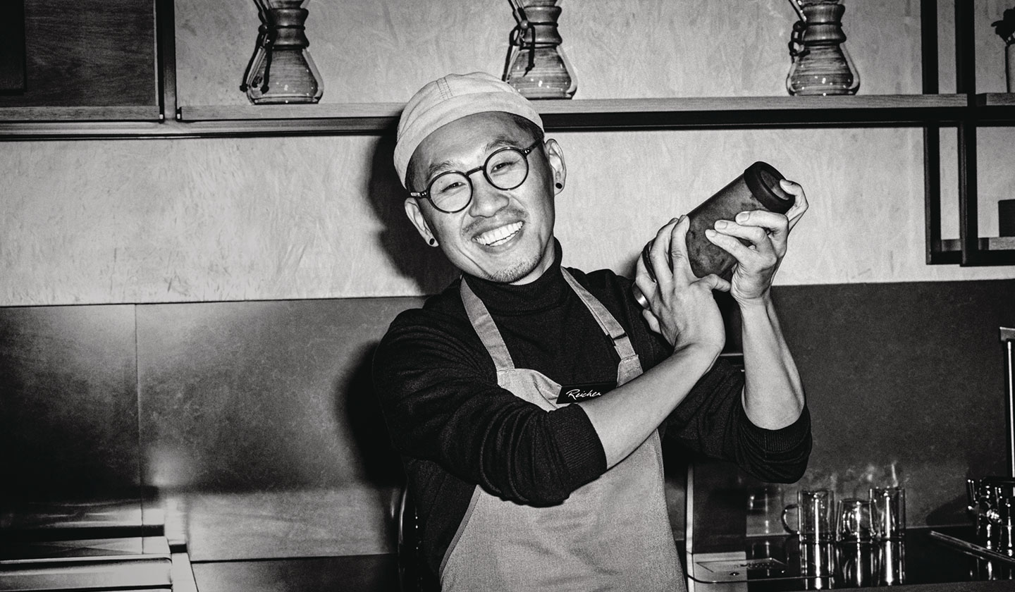 A friendly, welcoming barista shaking up an iced coffee with enthusiasm behind the counter.