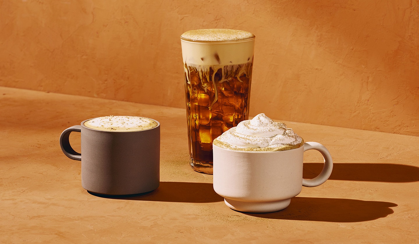 Three fall beverages sit against a warm orange background. On the left, a warm pecan oat latte is topped with golden pecan flavoured sugar in a grey mug. In the center, a deep brown iced pumpkin cream cold brew in a clear glass is layered with light beige cold foam and swirls of creamy white milk. On the right, a warm pumpkin spice latte in a white mug is topped with white whipped cream and a dusting of warm brown pumpkin pie spices.