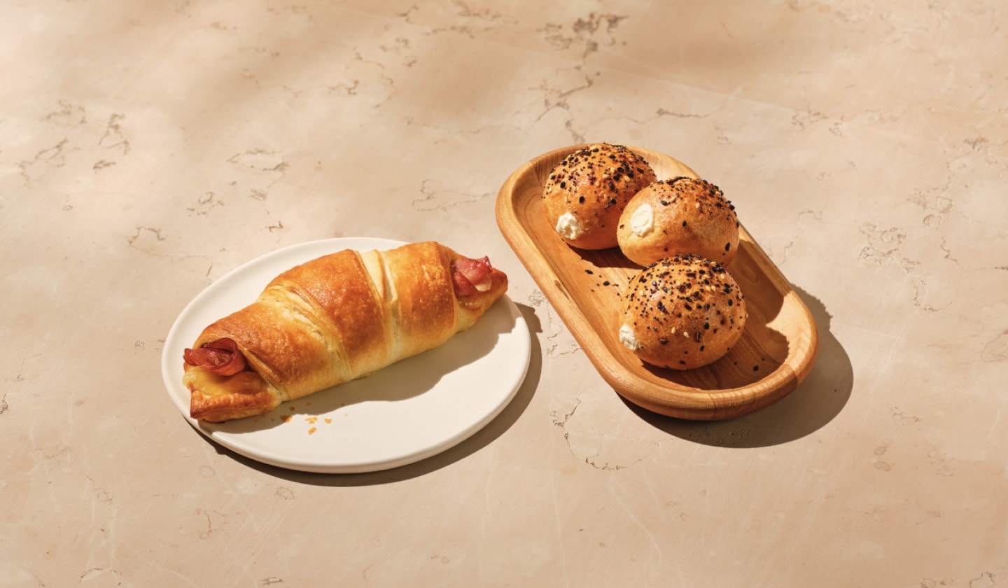2 savoury bakery snacks lie on a warm beige background. On the left, a freshly warmed flaky ham and swiss croissant. Next to it is a trio of golden mini bagel rounds topped with an assortment of seeds and seasoning.