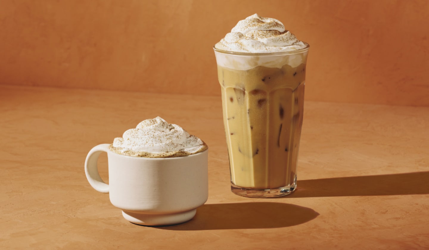 Un verre de latte glacé à la citrouille épicée beige et crémeux, chapeauté d’une riche mousse appétissante. À gauche, dans une tasse blanche, on voit la version chaude du latte, garni de crème fouettée attirante et d’épices pour tarte à la citrouille. Les deux boissons sont devant un chaleureux fond orangé.