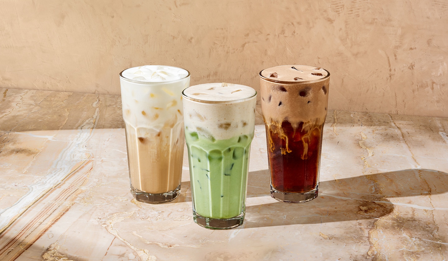 Nous voyons un latte glacé à la vanille rafraîchissant de couleur beige à gauche, à côté d’un matcha glacé à la banane vert laiteux au centre, et un café infusé à froid à la crème de chocolat brun riche à droite. Les trois boissons sont recouvertes d’une mousse protéinée crémeuse sur un fond beige chaud.