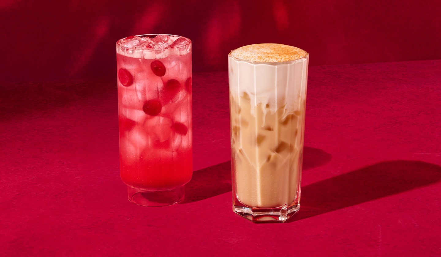 Une boisson glacée canneberge et orange aux couleurs éclatantes dans laquelle flotte des canneberges, à côté d’une boisson glacée chai crémeuse avec une garniture épicée, sur un fond rouge vif où se reflètent des lumières scintillantes.