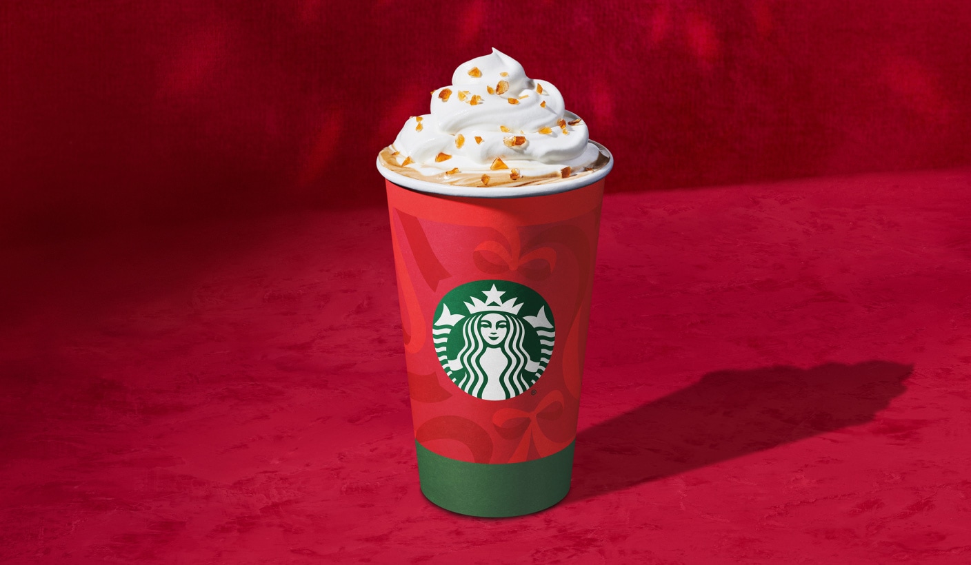 Une tasse des Fêtes remplie d’une boisson chaude à base d’espresso recouverte de crème fouettée et d’éclats de caramel croquants, sur fond rouge.