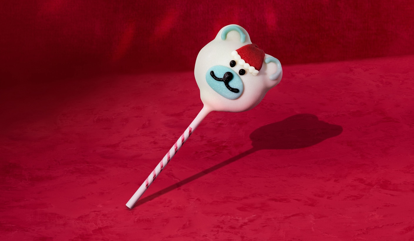 Un gâteau-suçon en forme d’ours sympathique sur un bâtonnet évoquant une canne en bonbon, sur fond rouge.