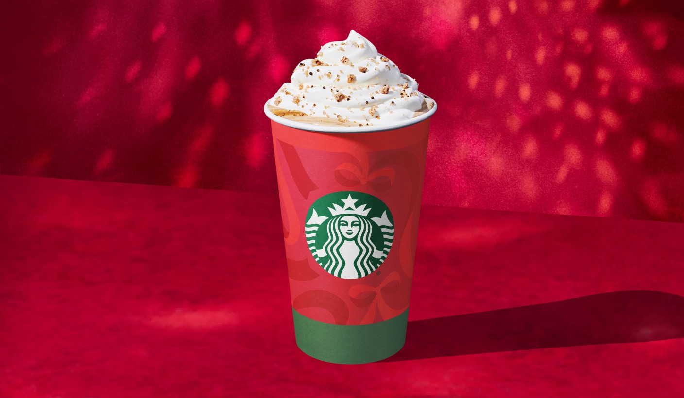 Photo d’un latte dans une tasse rouge des Fêtes dont le bas est orné d’une bordure verte. Nous voyons une couche de délicieuse crème fouettée saupoudrée d’une garniture de marron doré.