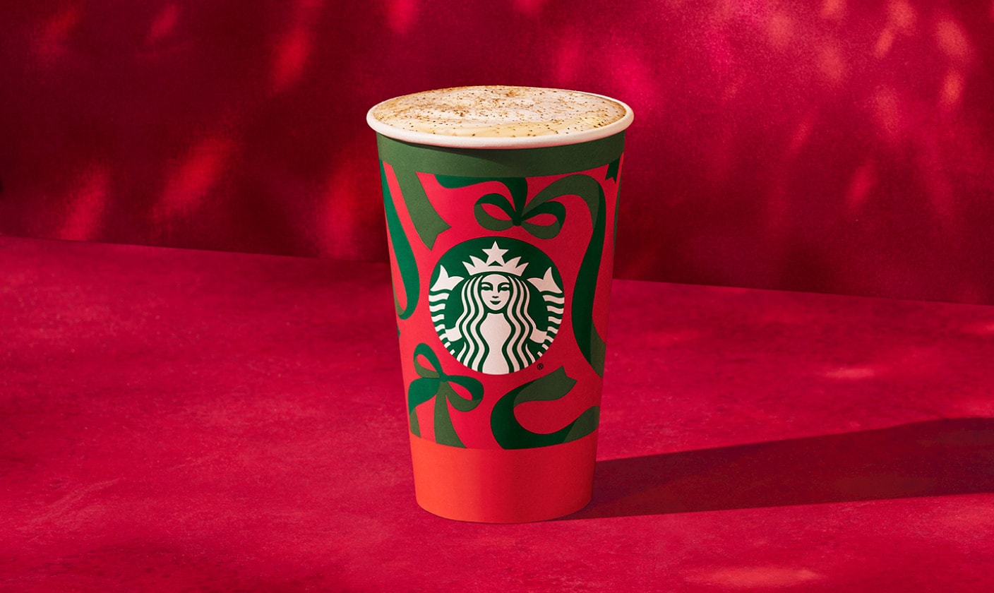 Photo d’un latte dans une tasse rouge des Fêtes ornée de boucles vertes, sur un fond en velours rouge vif où se reflètent des lumières scintillantes. La boisson est nappée de lait vapeur saupoudré de muscade dorée.