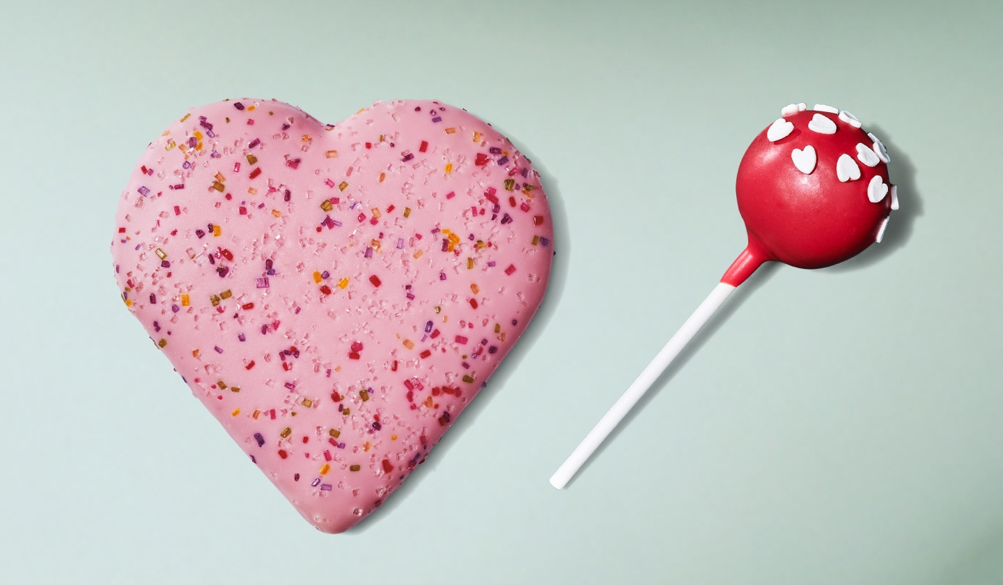 Un gros biscuit en forme de cœur, enrobé de glaçage rose et recouvert de paillettes colorées, à côté d’un gâteau-suçon rouge vif décoré de délicieux bonbons en forme de cœur, sur un fond vert pâle.
