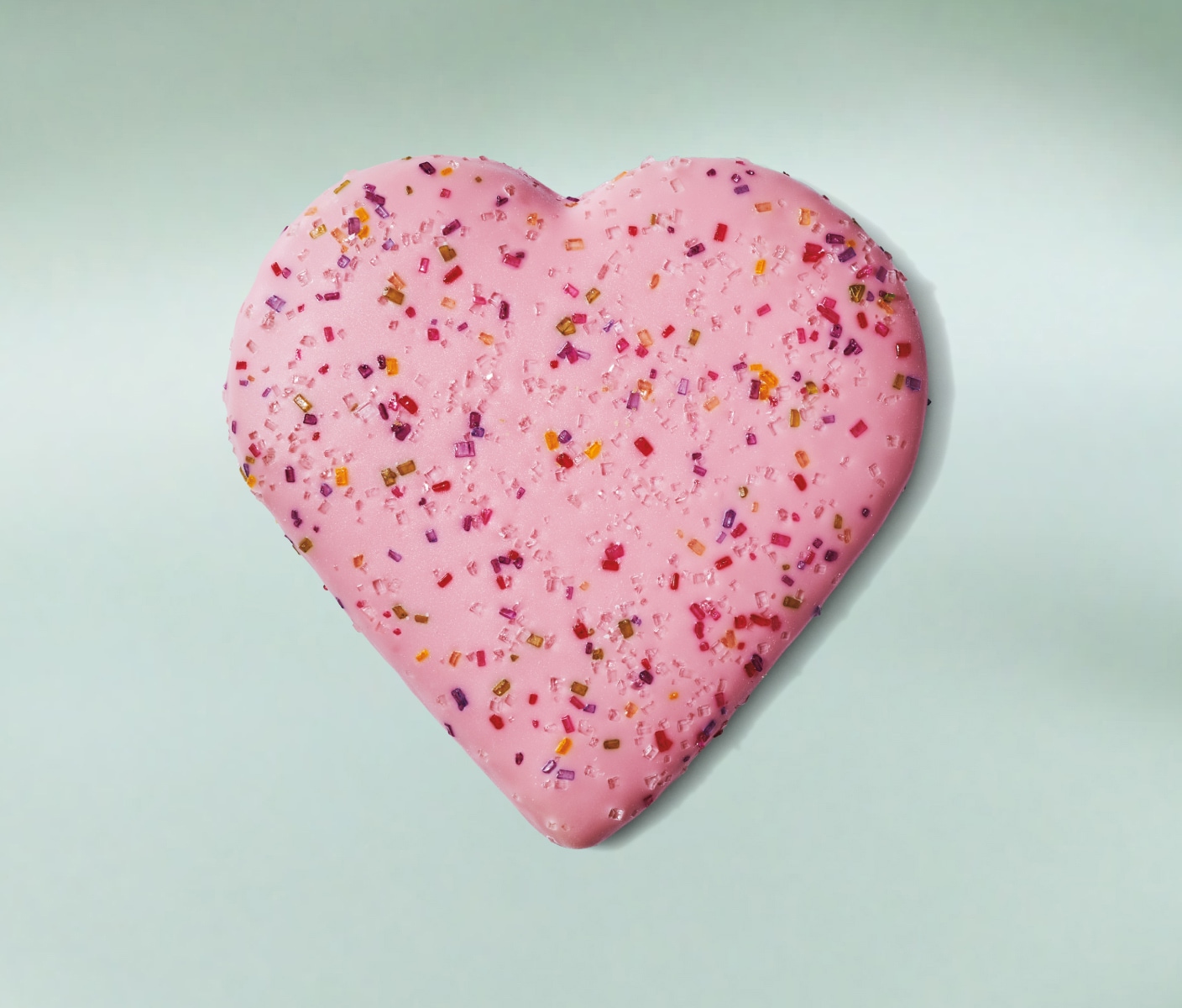 Un gros biscuit appétissant en forme de cœur, enrobé d’un épais glaçage rose et recouvert de paillettes colorées, sur un fond vert menthe.