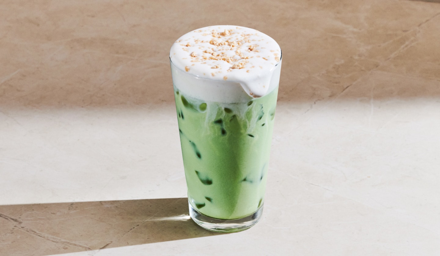 Un matcha glacé pain aux bananes est posé à la lumière du soleil. Une mousse froide recouverte de paillettes au caramel déborde sur le rebord du verre.