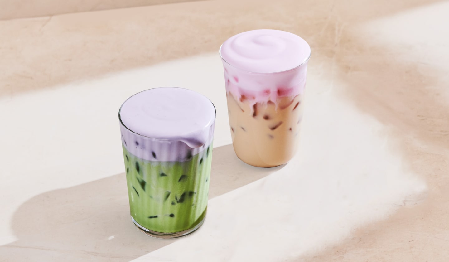 Deux boissons glacées sont superposées sur un fond neutre pâle. À gauche, on voit un matcha latte glacé vert coiffé d’une mousse froide violette; à droite, un chai latte glacé couronné d’une mousse froide à la cerise rose pâle.