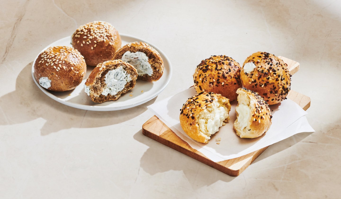 Des minibagels seigle et aneth sont posés sur une assiette, sur un fond neutre; l’un est coupé en deux pour montrer le fromage à la crème se trouvant à l’intérieur. À droite, des minibagels tout garnis sont posés sur un emballage, sur une planche à découper; ils sont ouverts, dévoilant une garniture de fromage à la crème.