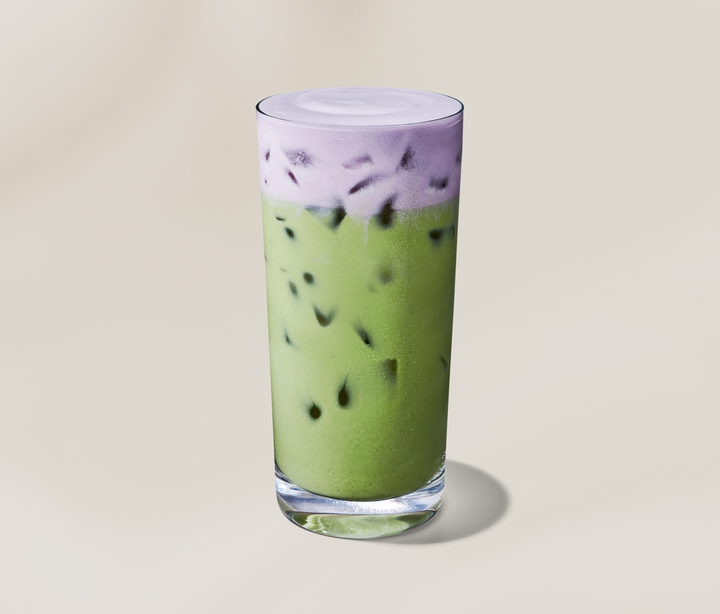 Un matcha latte glacé vert recouvert d’une mousse froide à la lavande de couleur violet dans un verre transparent, sur un fond beige.