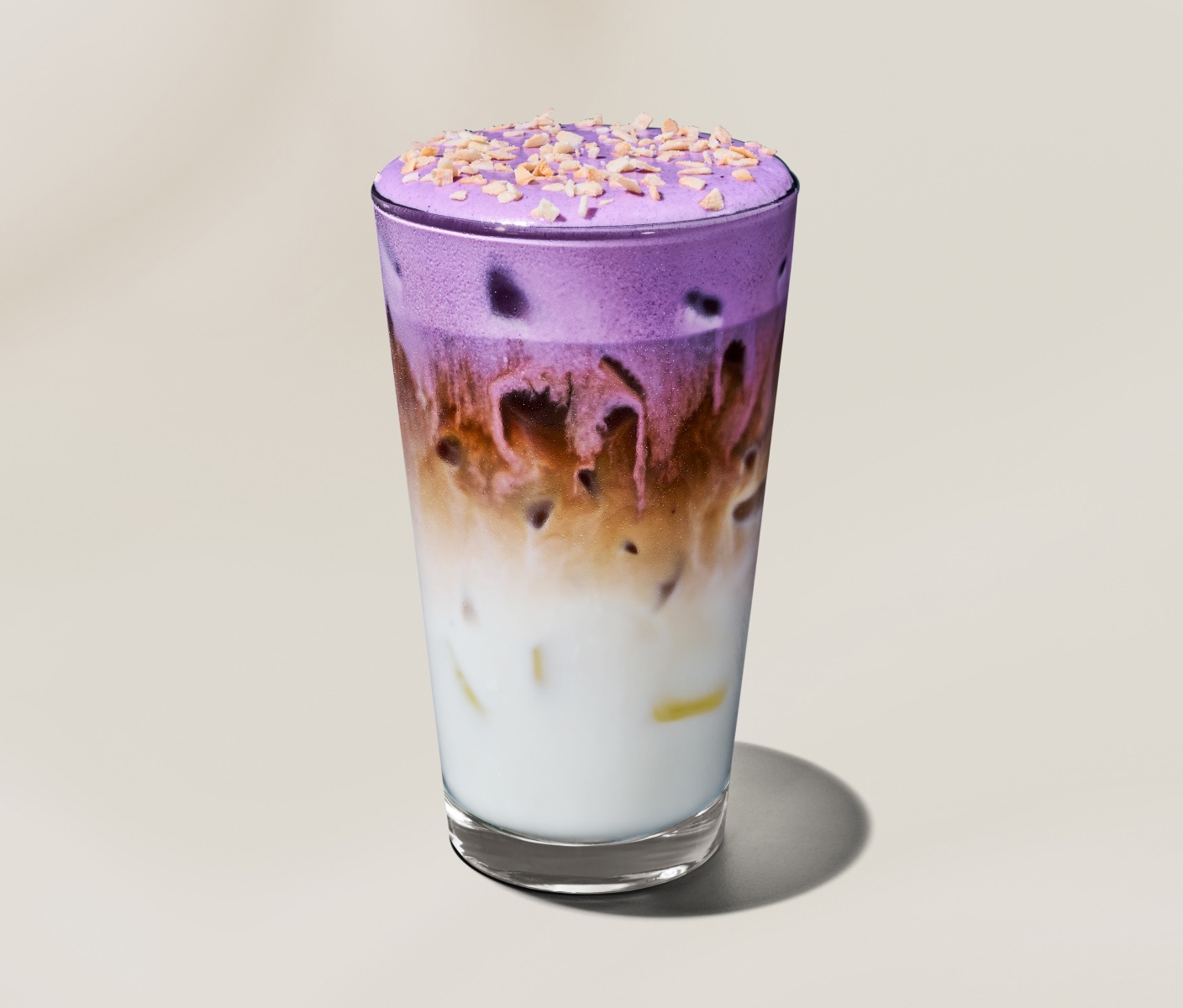 Un macchiato glacé ube et noix de coco dans lequel se superposent des couches d’espresso, de lait crémeux et de mousse froide violette à l’ube ainsi que des flocons de noix de coco, dans un verre transparent.