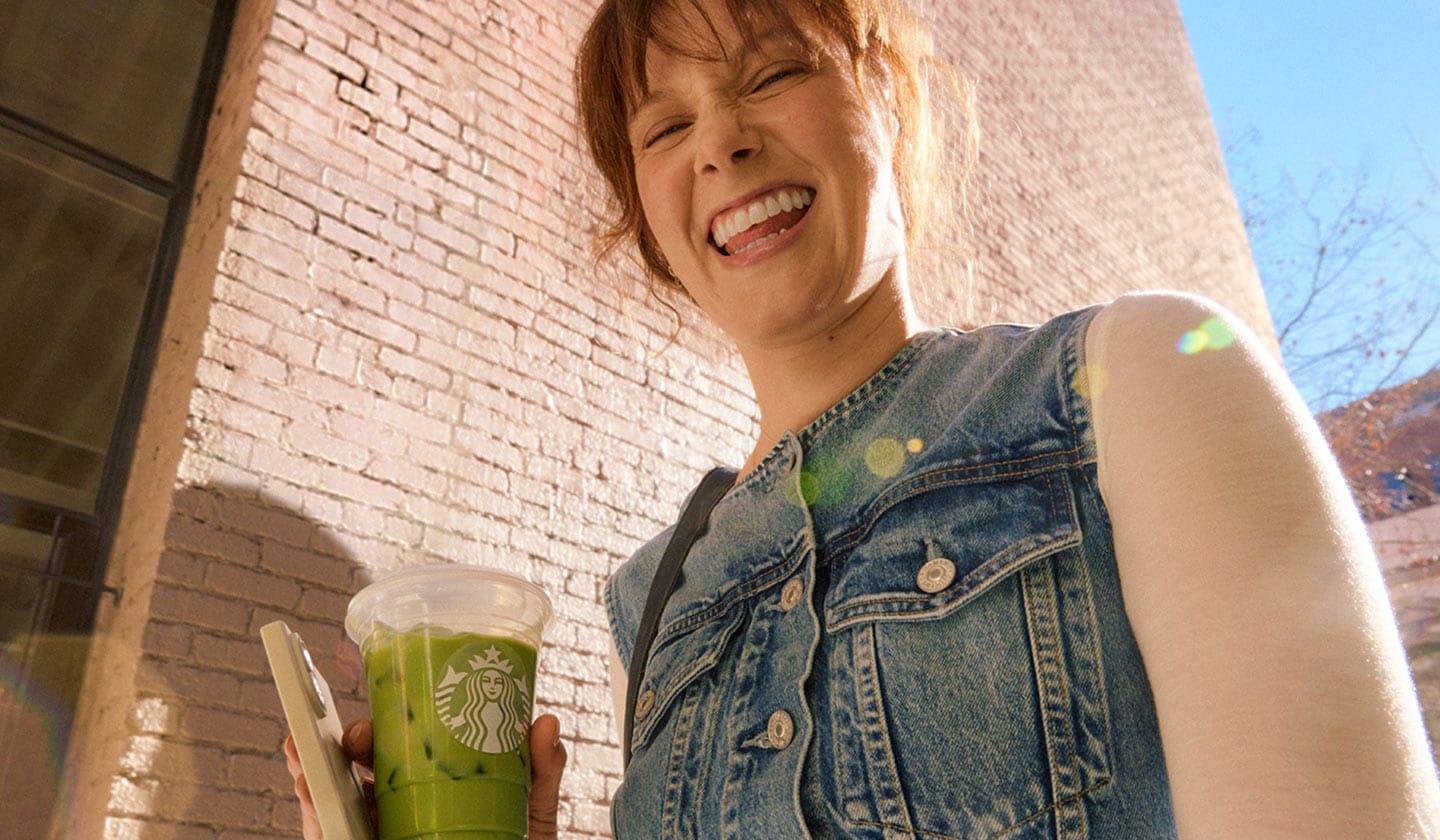 Une personne aux cheveux roux vêtue d’un gilet en jean tient une boisson glacée Starbucks verte et un téléphone devant un mur en briques.