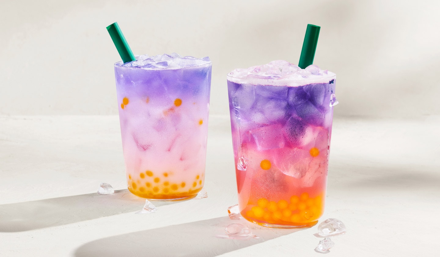 Deux boissons glacées rafraîchissantes aux trois couleurs distinctes (jaune éclatant, rose et violet) sur fond gris pâle, chacune avec une paille verte.