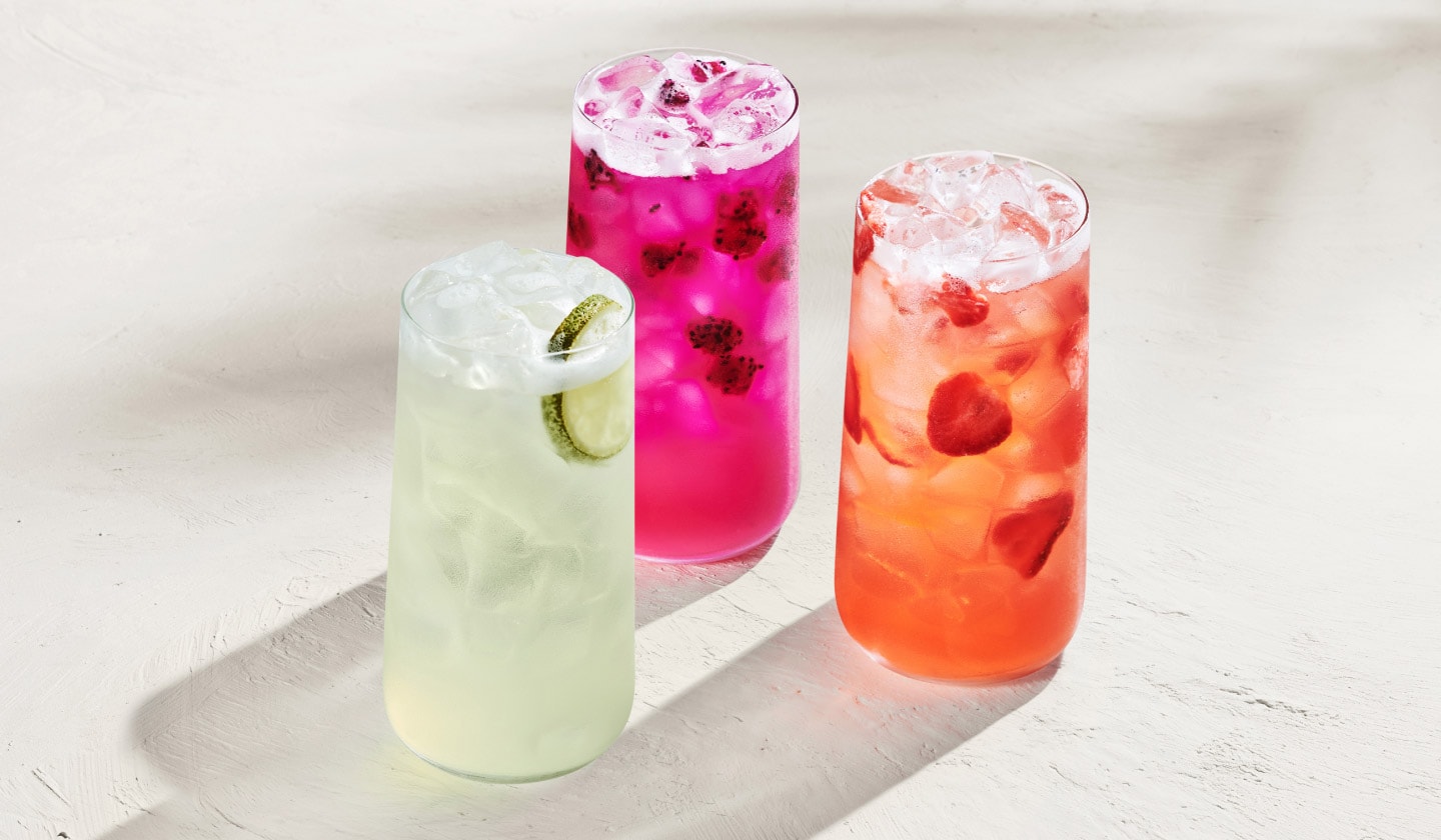 Une boisson glacée vert pâle garnie d’une tranche de lime, une boisson glacée rouge clair avec des morceaux de fraise et une boisson glacée rose vif au fruit du dragon, présentées sur un fond blanc lumineux.