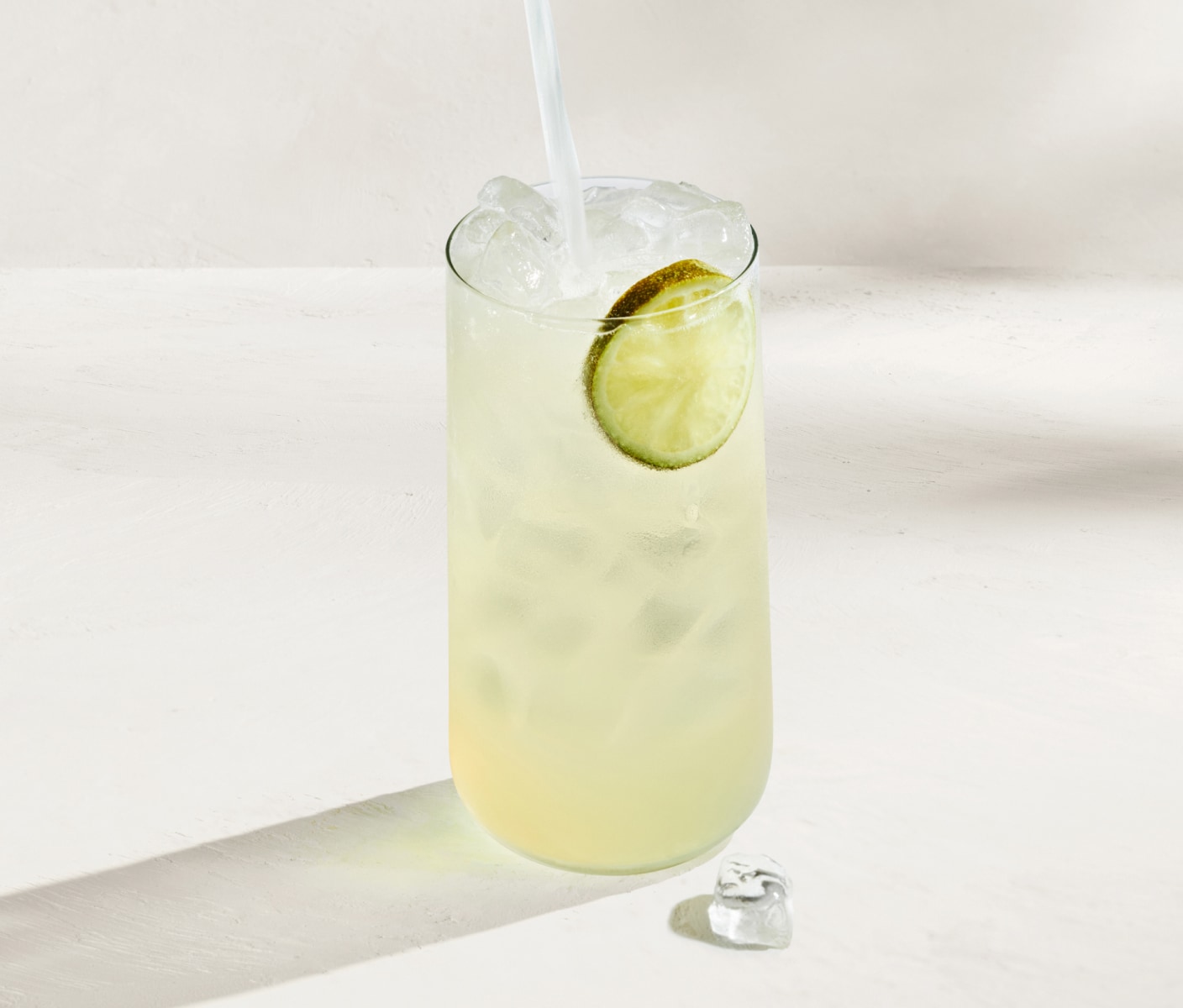 Boisson vert pâle, glacée et rafraîchissante, garnie d’une tranche de lime, sur un fond blanc baigné de soleil.