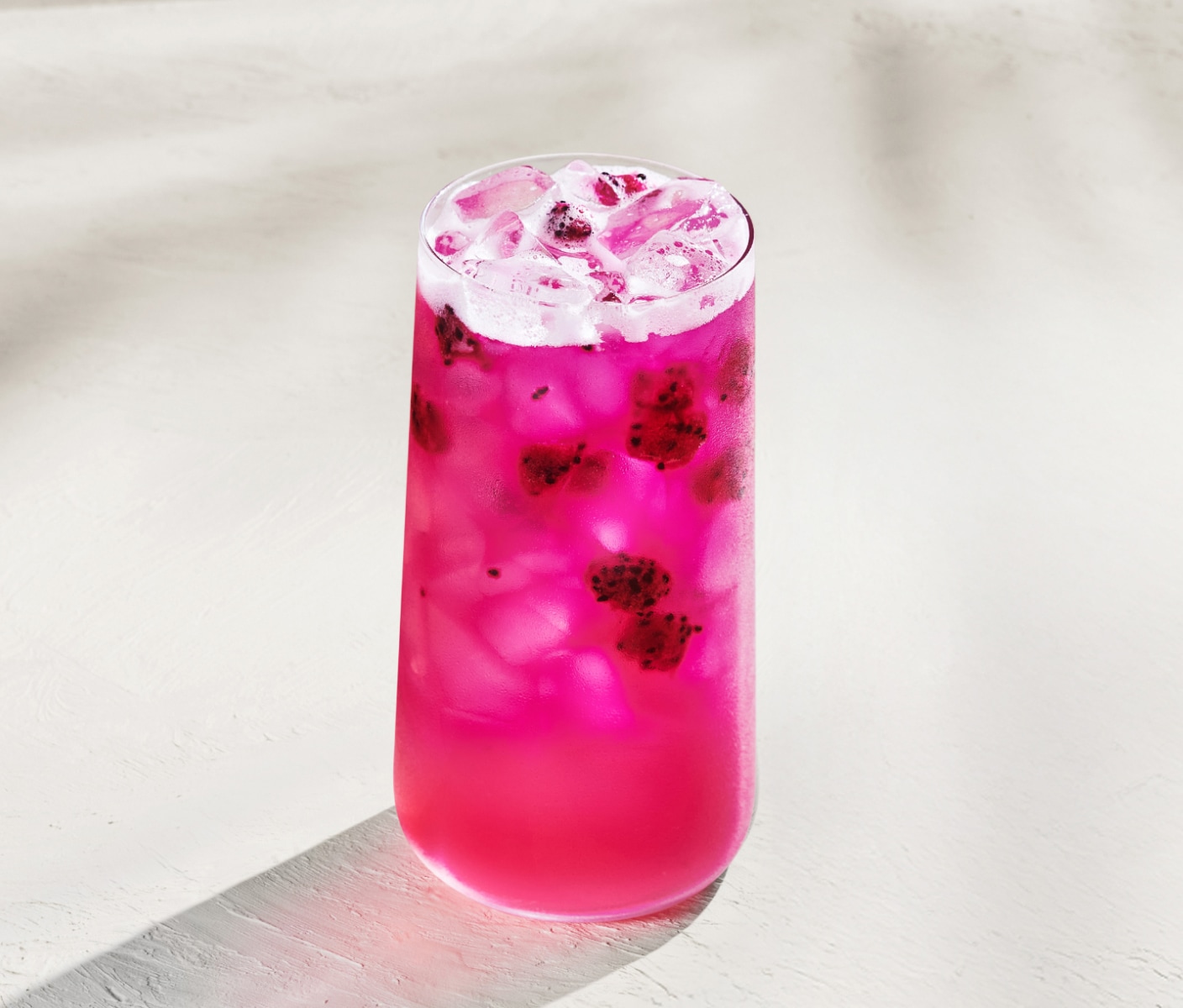 Un verre de boisson glacée rose garnie de morceaux de fruit du dragon, présenté sur un fond beige.