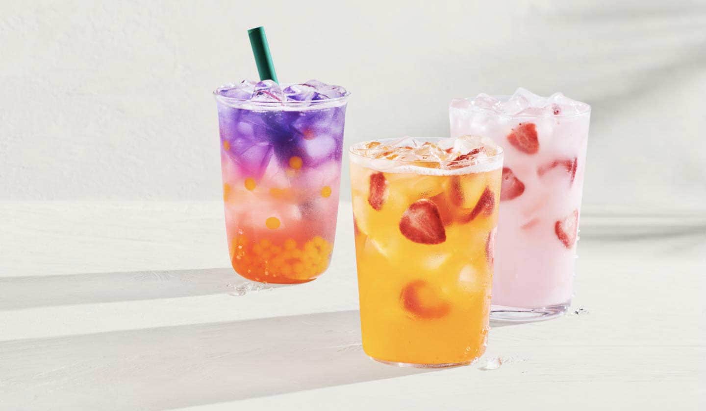 Trois boissons rafraîchissantes sont présentées sur un fond blanc lumineux. À gauche, on voit une boisson glacée aux saveurs tropicales : perles dorées aromatisées à la mangue et à l’ananas, goyave rose et infusion de fleur de pois papillon violette. Au centre, on voit une boisson glacée d’un jaune doré à saveur de mangue dans laquelle flottent des morceaux de fraises séchées. À droite, on voit une boisson à la fraise et à la noix de coco, aussi agrémentée de morceaux de fraises séchées.