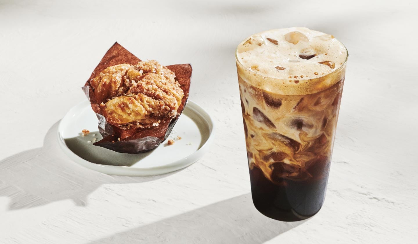 On voit une pâtisserie à la cannelle enveloppée de papier brun et posée sur une assiette blanche ainsi qu’une boisson glacée à base d’espresso, toutes deux sur un fond blanc lumineux.