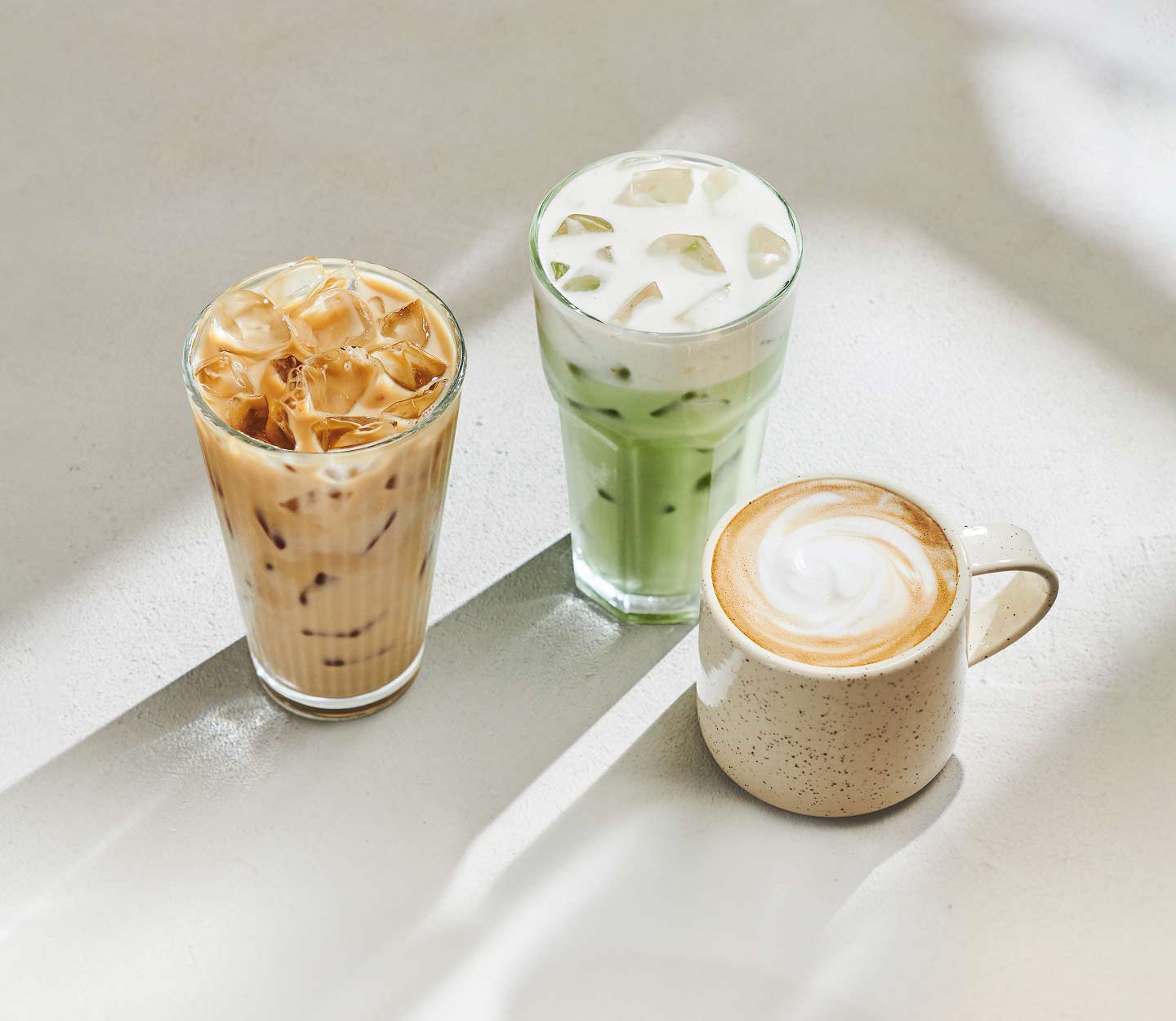 Trois boissons protéinées disposées sur une table pâle, dont un latte glacé, une boisson glacée verte et un latte chaud avec un motif dans la mousse.