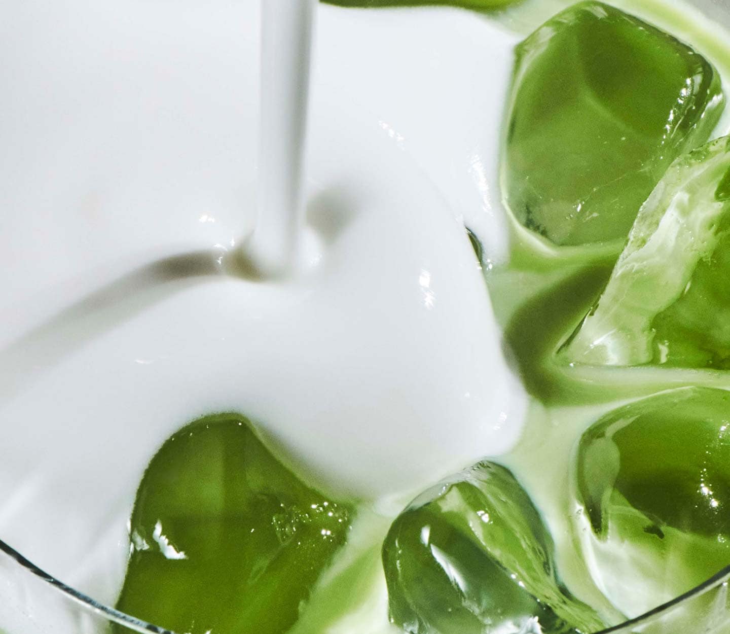 Gros plan sur du lait versé dans une boisson glacée verte, tournoyant autour de gros glaçons, dans un verre.