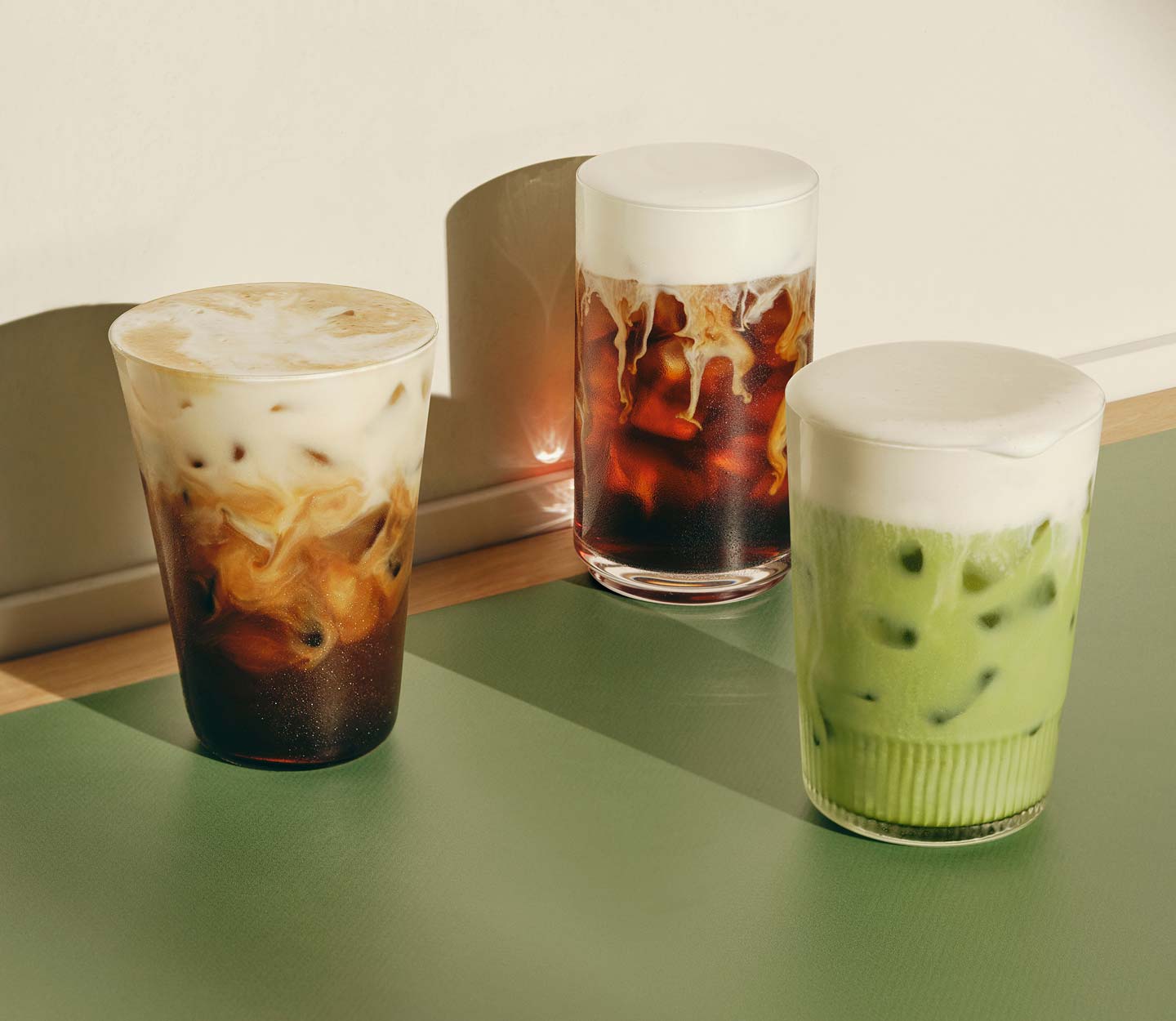 Trois boissons glacées maison sur une table, dont une boisson glacée verte et deux boissons foncées à base de café garnies d’une mousse protéinée crémeuse.