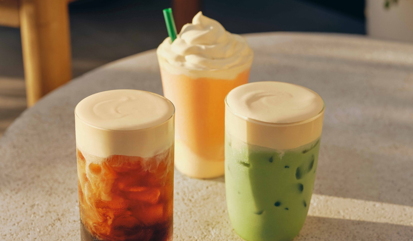 Trois boissons glacées dans des verres sont posées sur une table en béton, à l’extérieur. À gauche, on voit un café glacé coiffé d’une mousse froide à l’orange; au centre, une boisson frappée orange coiffée d’une mousse froide à l’orange; et à droite, un matcha glacé coiffé d’une mousse froide à l’orange.