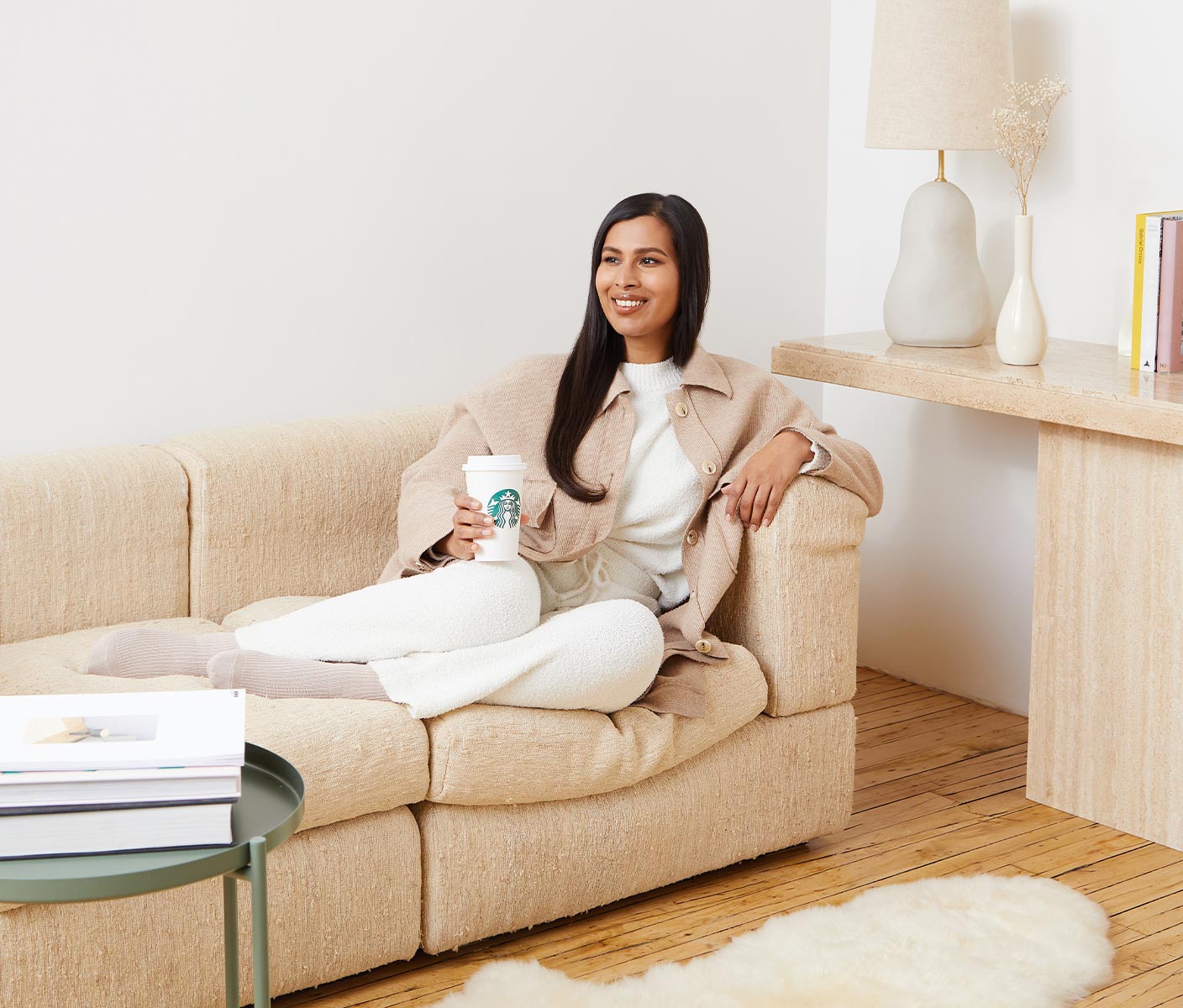 Une jeune femme assise dans un canapé dans sa demeure chaleureuse et lumineuse tient une commande Starbucks en souriant avec optimisme.