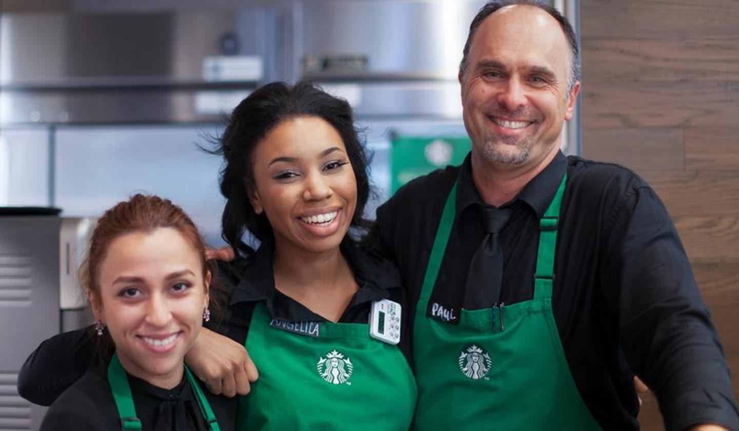 Trois partenaires (employés) de Starbucks portant un tablier vert qui se tiennent debout côte à côte en souriant.