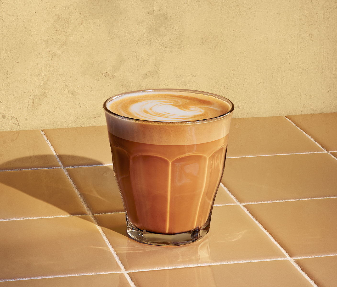 Une boisson espresso dorée couronnée d’un tourbillon de mousse de lait. Servie dans un verre posé au soleil sur un comptoir en carrelage.