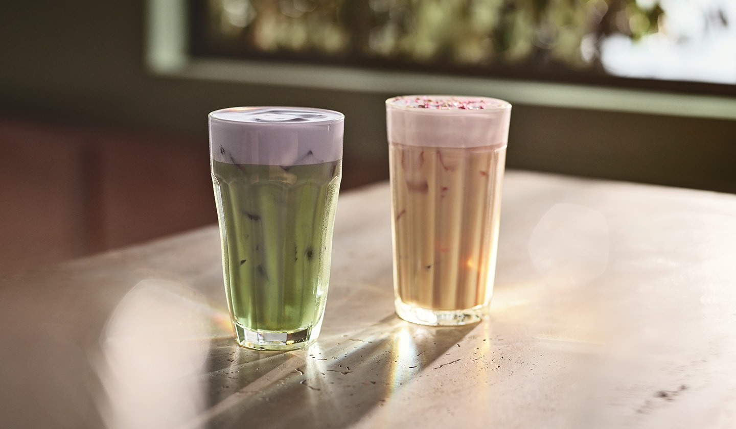 Le soleil éclaire une table où se trouvent matcha vert glacé garni de mousse froide violette et un chai glacé garni de mousse froide et de paillettes roses.