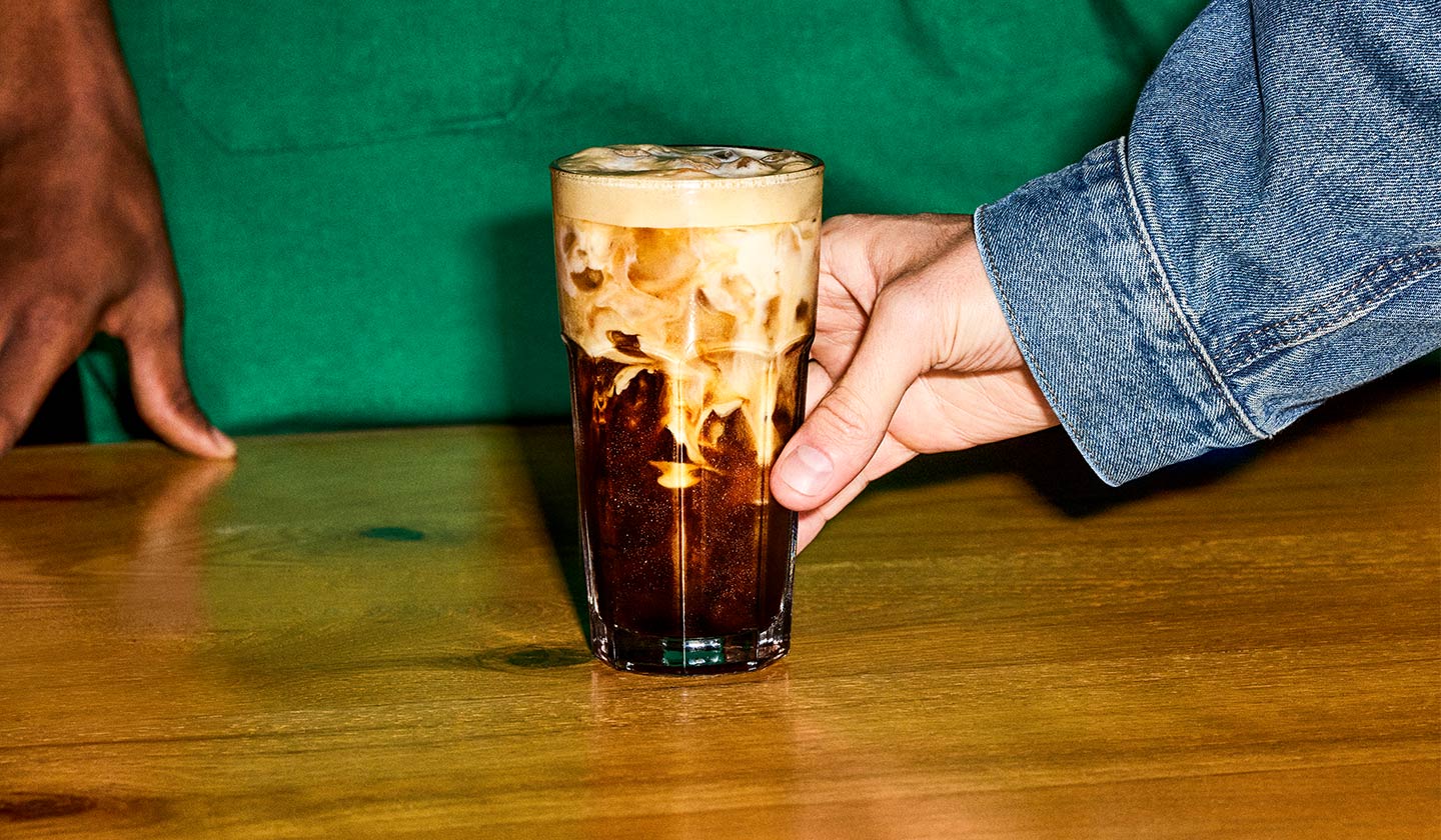 Un client tend le bras pour prendre une boisson espresso artisanale à déguster sur place. Un barista se tient derrière le comptoir.