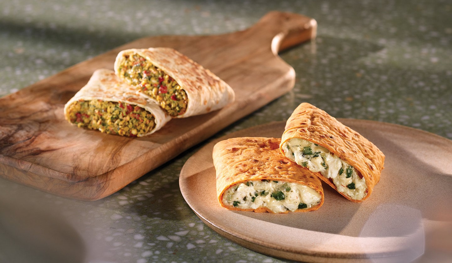Deux savoureux wraps posés sur des planches en bois et sur lesquels la lumière du soleil projette des ombres légères. L’un est rempli de falafel et d'herbes, l’autre de poulet, de jalapeño et de fromage.