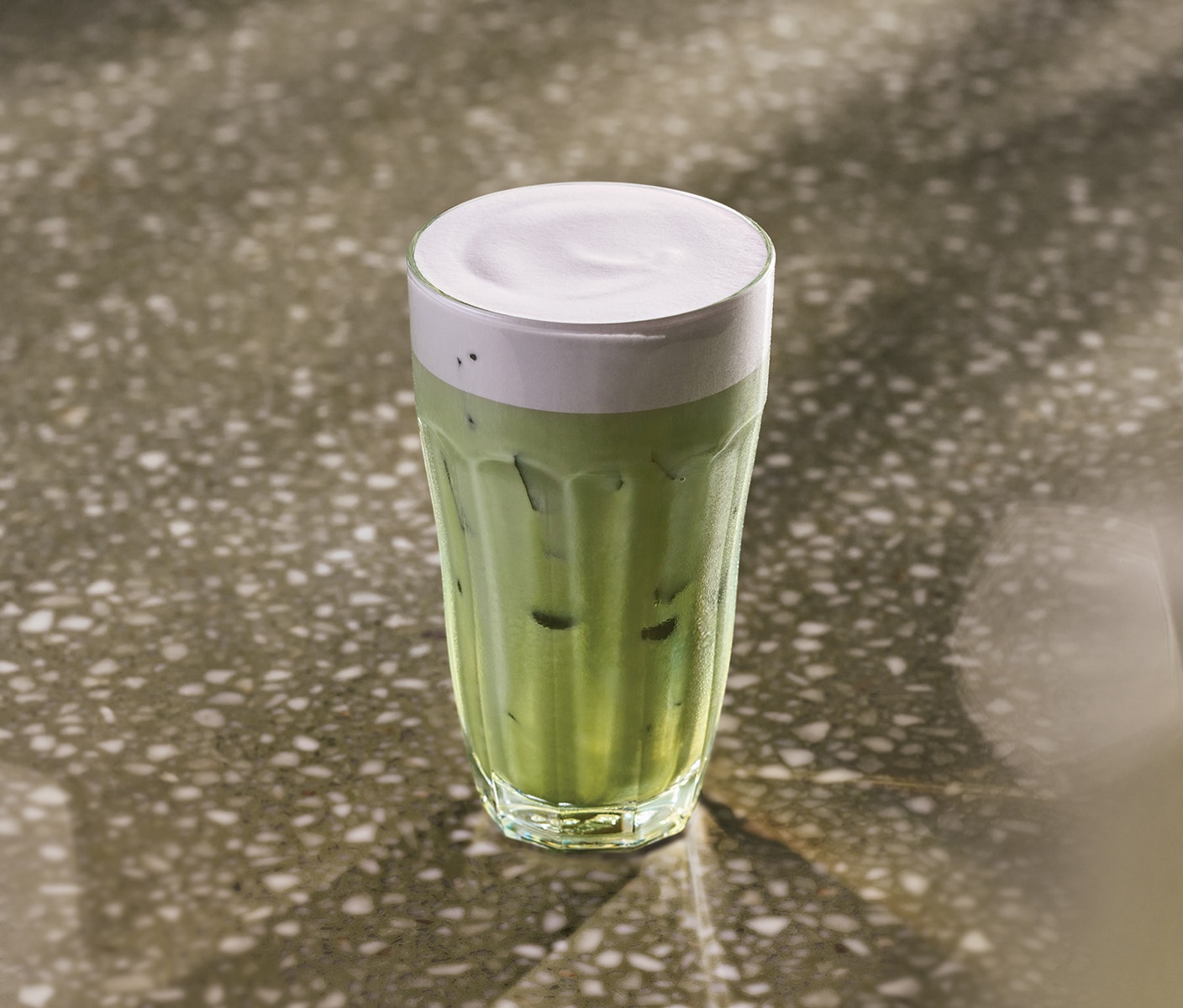 Le soleil éclaire une table où se trouve une boisson glacée au matcha vert couronnée de mousse froide violette, servie dans un verre.