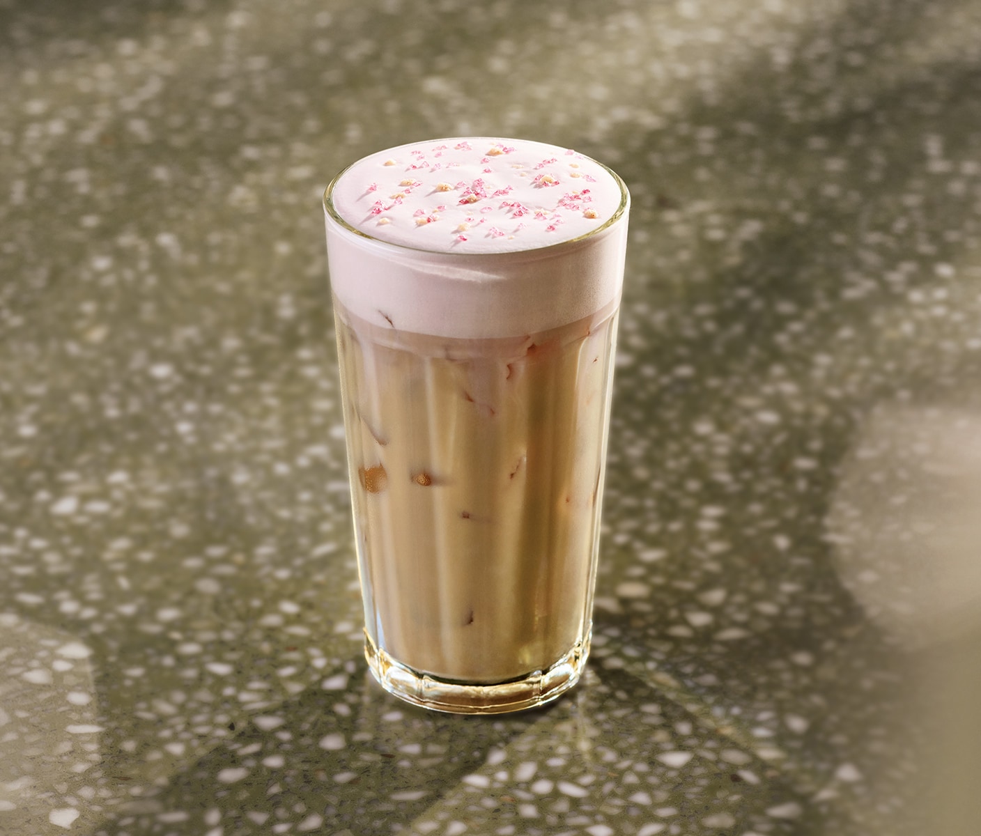 Le soleil éclaire une table où se trouve une boisson glacée au chai couronnée de mousse froide rose et d’une garniture croquante rose, servie dans un verre.