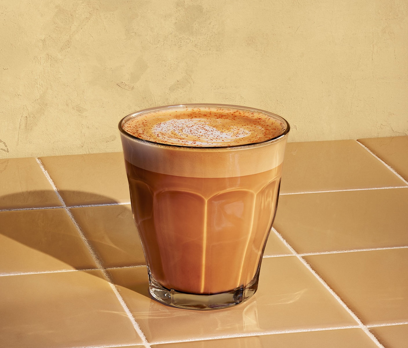 Une boisson espresso dorée couronnée d’un tourbillon de mousse de lait, avec une pincée de cannelle sur le dessus. Servie dans un verre posé au soleil sur un comptoir en carrelage.
