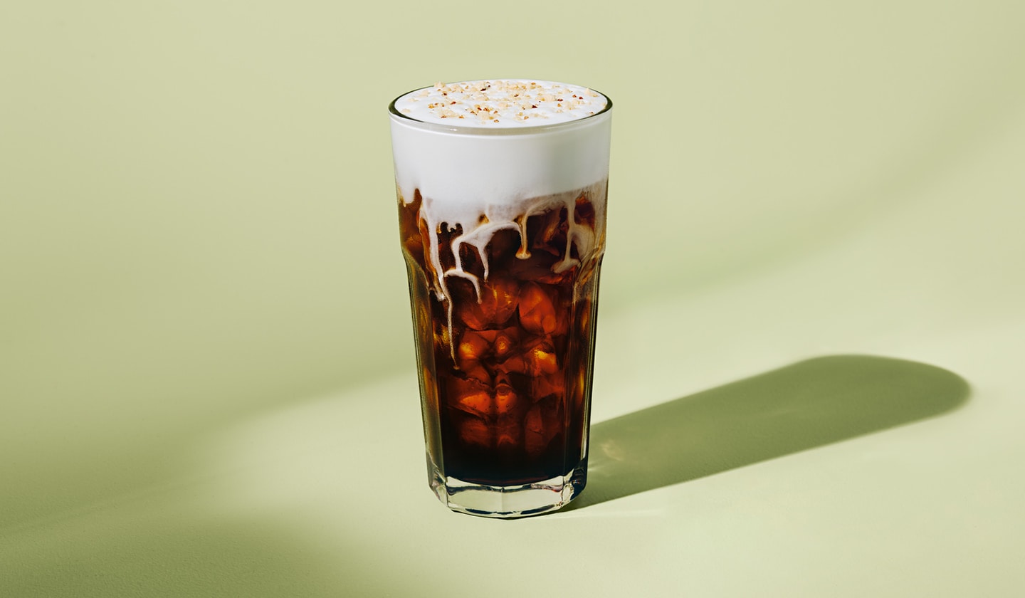 Un rafraîchissant café infusé à froid couronné d’une mousse froide crémeuse garnie de miettes de biscuits grillés, sur fond vert clair.
