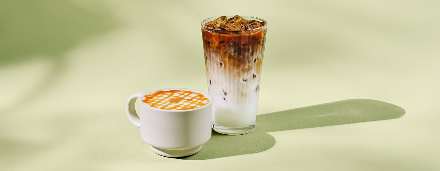 Un macchiato chaud avec filet de caramel dans une tasse blanche à côté d’un macchiato glacé dans un grand verre, sur fond vert clair, avec ombres légères.