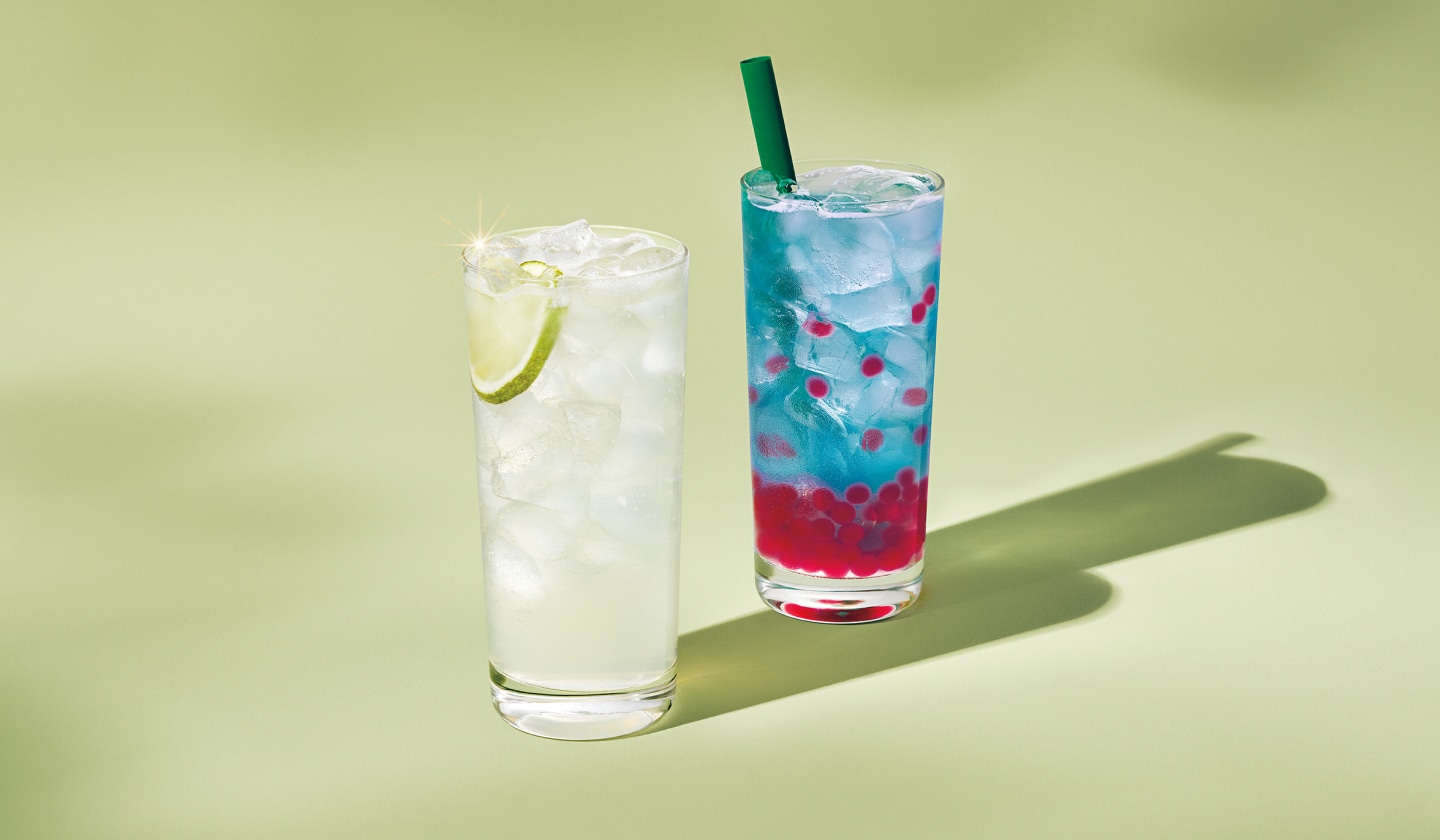 Un fond vert doux avec deux verres. Le premier est translucide avec de la glace et une tranche de citron vert. L'autre est bleu avec de la glace et des perles de couleur rouge vif.