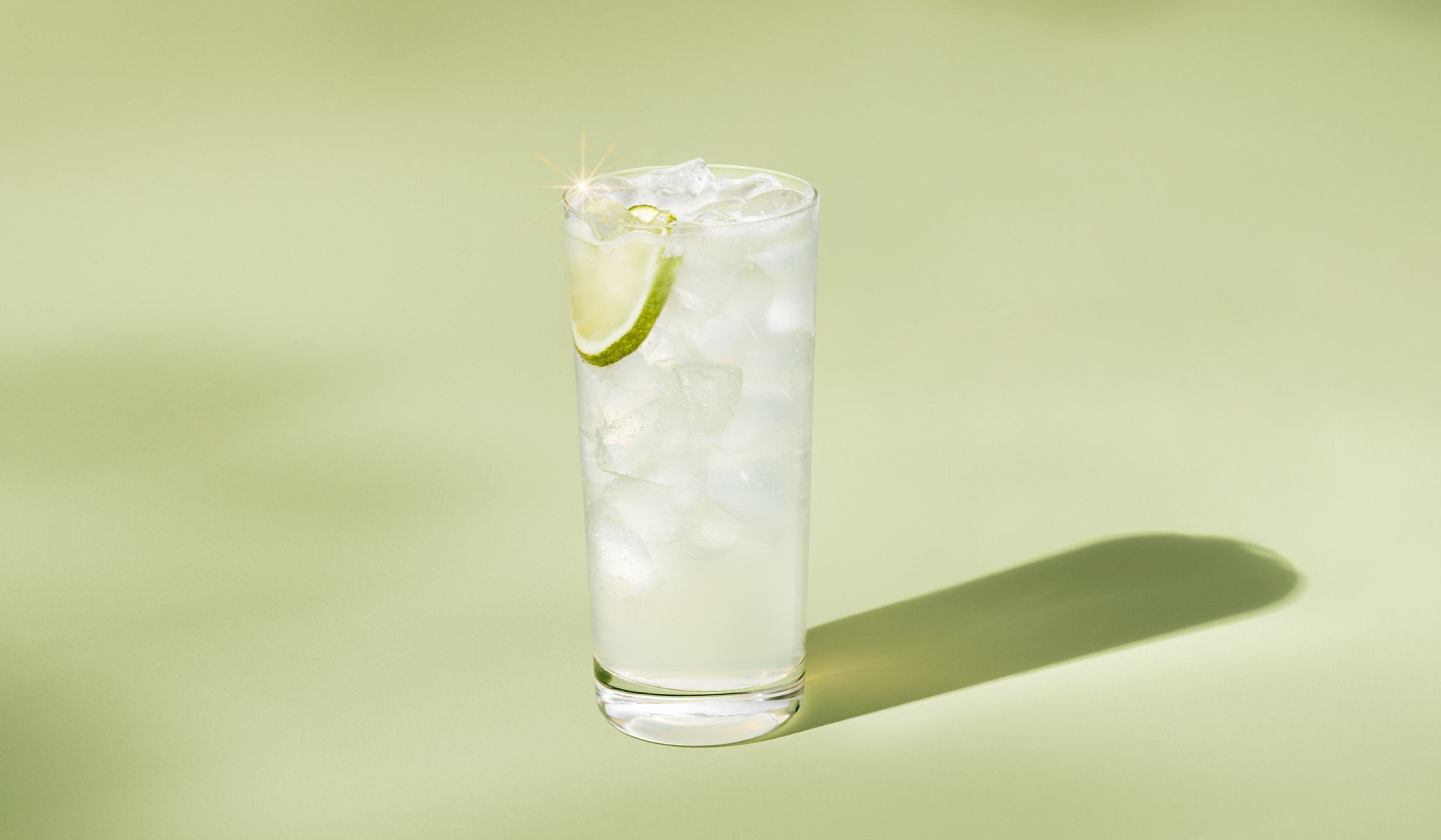 Un grand verre contenant une limonade translucide remplie de glace. Une tranche de lime est suspendue dans la glace, sur fond vert doux.