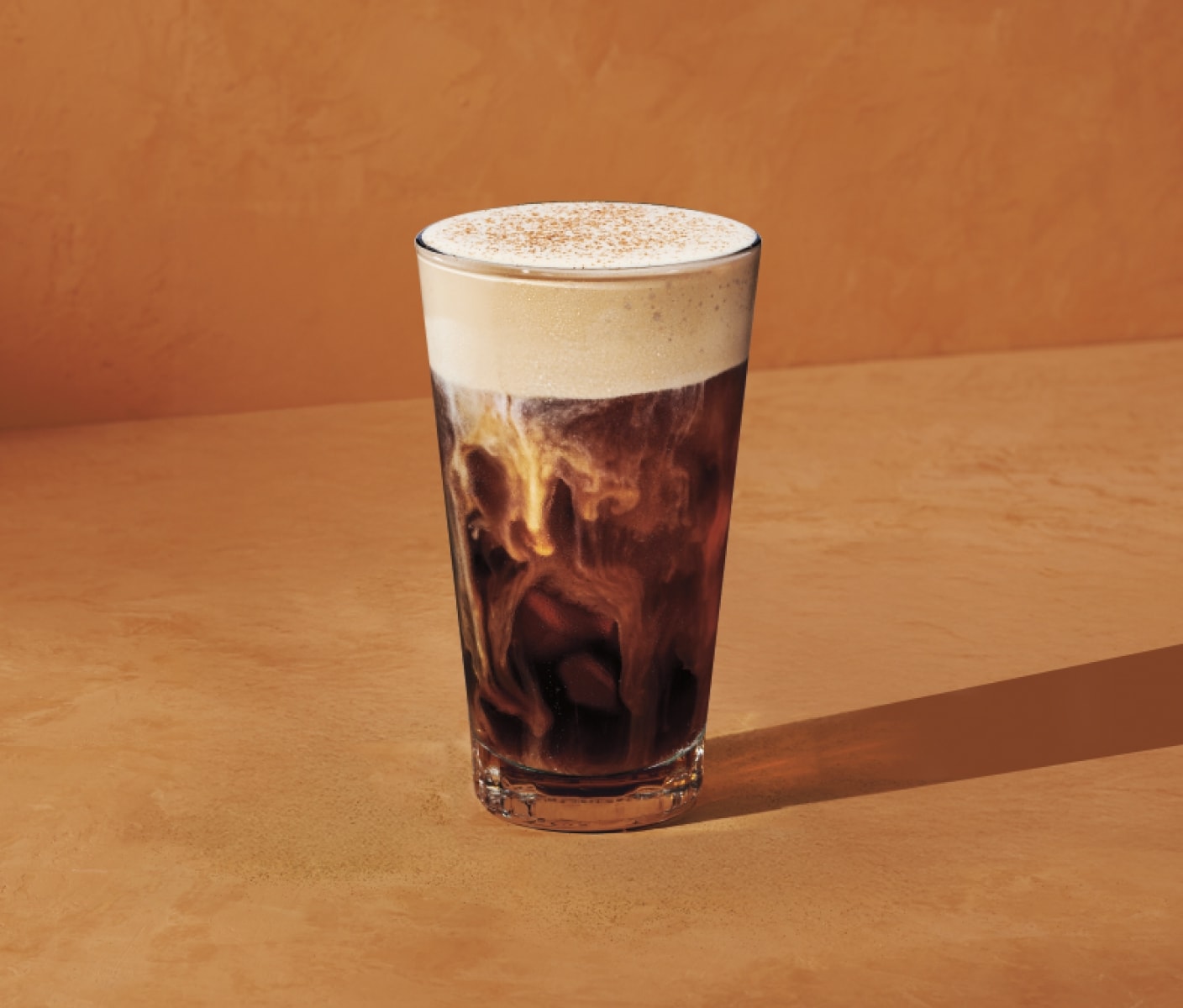 Sur un fond orange chaleureux, on voit, dans un verre transparent, un café infusé à froid avec crème à la citrouille, garni d'une mousse pâle sous laquelle on aperçoit des tourbillons de lait blanc crémeux.