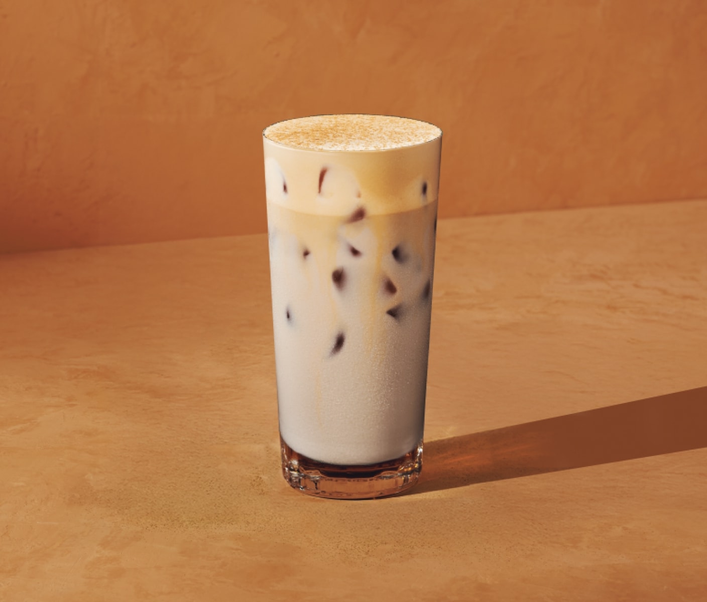 Posé sur un fond orange chaleureux, on voit un chai latte glacé riche et onctueux beige, recouvert d’une mousse délicieuse et saupoudré d’une garniture à la citrouille épicée.