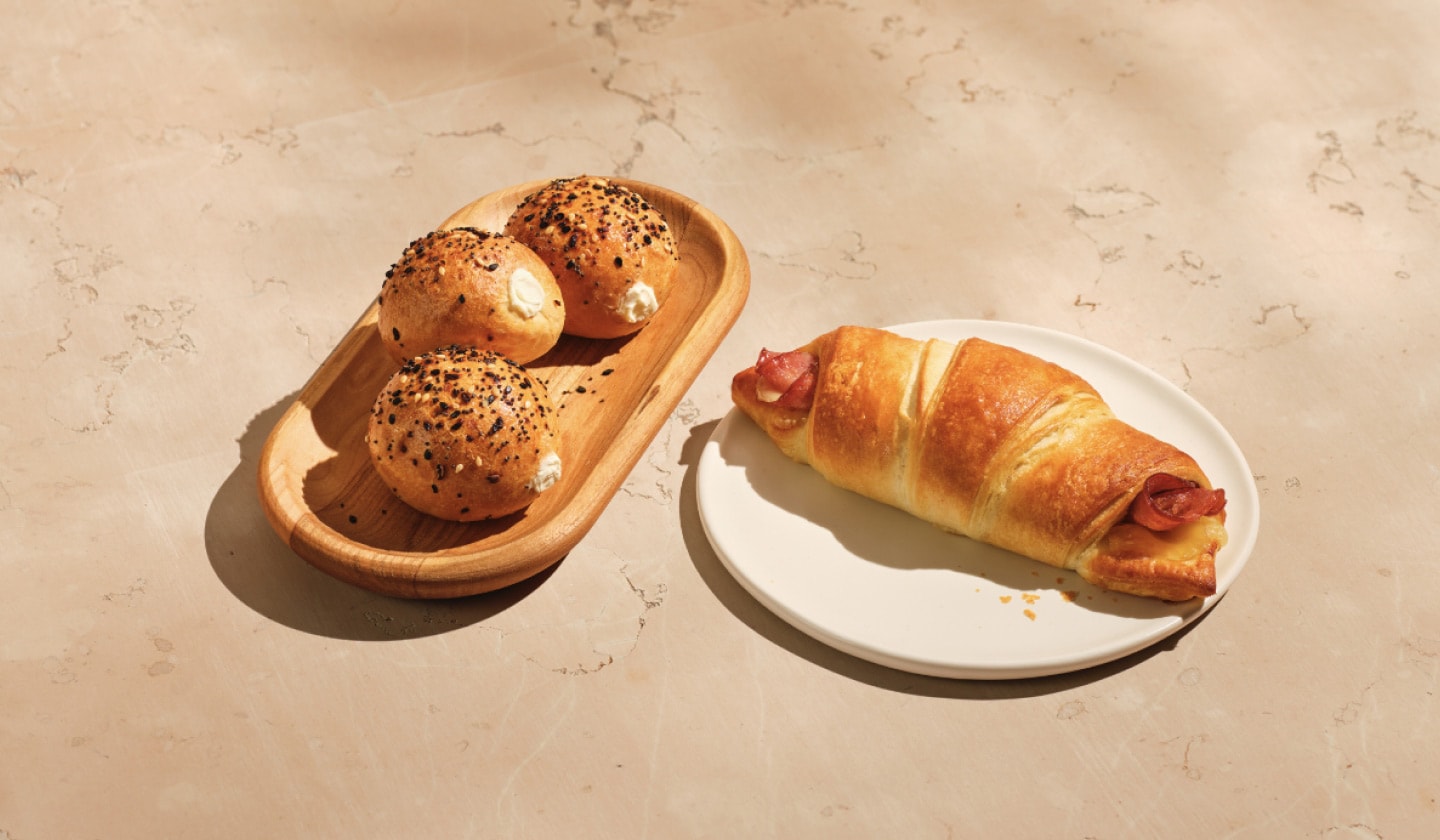 Deux produits de boulangerie sont présentés sur un doux fond beige. À gauche, un trio de minibagels dorés garnis d'une variété de graines et d'assaisonnements. À droite, le croissant feuilleté chaud au jambon et au fromage suisse.